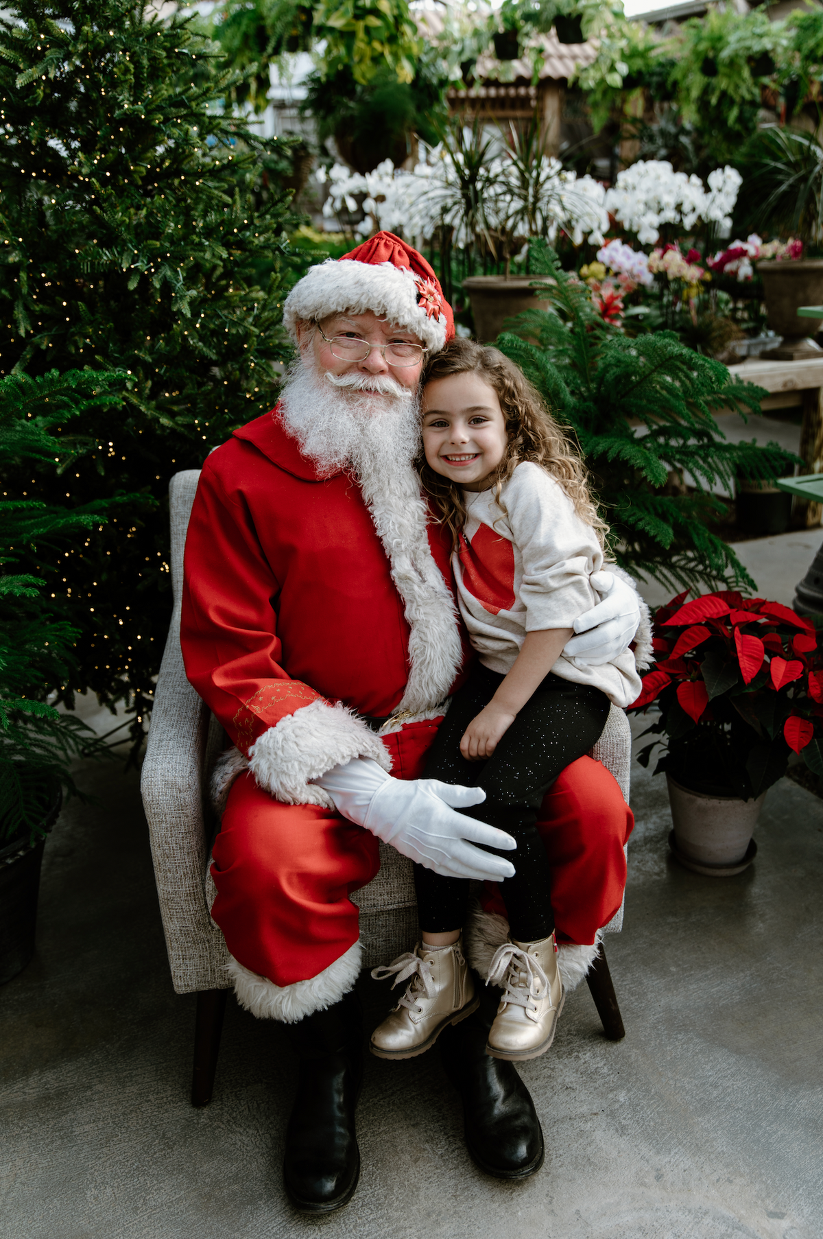 A young girl sitting on Santa Claus's lap for a photograph in a greenhouse setting with Christmas decorations.